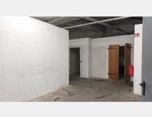 Ein Raum mit weißen Ziegelwänden und einem Betonboden, teilweise geöffneten Holztüren auf der rechten Seite und freiliegenden Rohren an der Decke. An der Wand hängt ein roter Feuerlöscher.