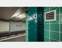 U-Bahn-Station mit einem Bereich der Haltestelle "Wittenbergplatz". Zu sehen sind grün geflieste Wände, eine genietete grüne Metallstütze, ein Lüftungsgitter und eine Wand aus weißen Kacheln mit einem Schild, das den Stationsnamen trägt.