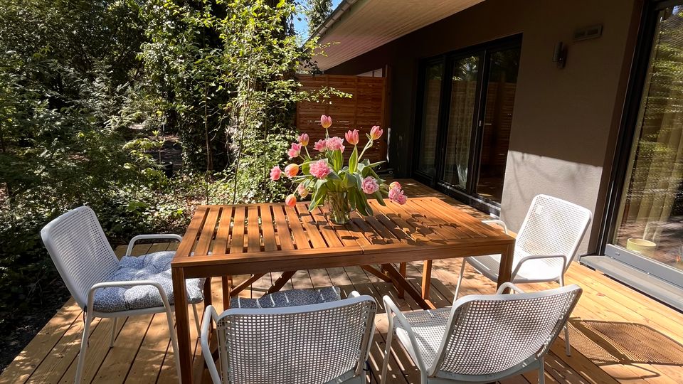 Eine Terrasse mit Holzboden, darauf ein rechteckiger Holztisch mit vier weißen Stühlen und einem Blumenstrauß in der Mitte. Im Hintergrund sind eine Hausfassade mit Glasfenstern und ein dicht bewachsener Garten mit Bäumen zu sehen.