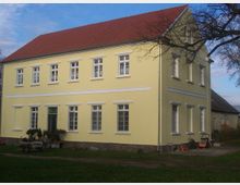 Zweistöckiges, hellgelbes Wohngebäude mit Satteldach aus roten Dachziegeln, zahlreichen weißen Fenstern und einem steinernen Sockel. Im Vordergrund befindet sich ein Hof mit Rasenfläche und Gartenmöbeln.