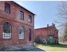 Zwei historische Backsteingebäude mit großen, bogenförmigen Fenstern; das linke Gebäude hat rote Holzdetails an Türen und Fenstern, und das rechte zeigt eine grüne Plane mit einem Tiermotiv. Der Bereich ist von Gras und Bäumen umgeben.