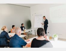 Ein heller Seminarraum mit fünf sitzenden Teilnehmern an weißen Tischen, die einer Präsentation folgen. Eine Vortragende in Businesskleidung steht neben einem Flipchart mit Diagramm und erklärt Inhalte, während im Hintergrund eine Projektion auf der Wand sichtbar ist.