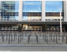 Die Vorderseite der Mercedes-Benz Arena mit einer modernen Glasfassade und einem Vordach, dahinter ein großer Schriftzug mit dem Arenanamen. Im Vordergrund befinden sich zahlreiche Metallgeländer, die den Eingangsbereich strukturieren.