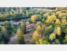 Luftaufnahme eines Hauses mit rotem Ziegeldach, eingebettet in eine bewaldete Landschaft. Im Hintergrund fließt ein Fluss, umgeben von dichten Bäumen, und ein schmaler Weg verläuft parallel zum Ufer.