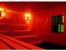 Holzvertäfelter Sauna-Innenraum mit mehreren Sitz- und Liegebänken aus Holz, warmem orangefarbenem Licht an den Wänden und beheizten Elementen. Zwei grün leuchtende Paneele sind an der rechten Wand angebracht.