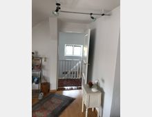 Heller Innenraum eines Wohnhauses mit weiß gestrichenen Wänden und Holzboden. Ein kleiner Beistelltisch mit Lampe steht rechts, während eine offene Tür einen Blick auf einen Flur mit Treppe und einem Fenster im Hintergrund gewährt, ergänzt durch Teppiche auf dem Boden.