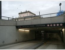 Einfahrt zu einem Tiefgaragenparkplatz mit der Beschriftung „Q-Park Unter den Linden/Staatsoper“. Im Hintergrund ist ein historisches Gebäude mit Statuen zu sehen, der Himmel ist bewölkt.