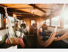 Innenraum eines Holzbootes mit mehreren Menschen, die auf Hängematten und Bänken sitzen oder liegen. Große Fenster lassen Sonnenlicht herein, das den Raum warm beleuchtet.