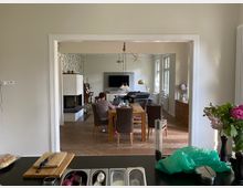 Blick von einer Küche in ein Wohnzimmer mit Essbereich. Im Vordergrund eine Küchenanrichte mit Schneidebrett und Gemüse, dahinter ein Holztisch mit Stühlen, ein Kamin, ein Sofa, ein Fernseher an der Wand und große Fenster, die viel Tageslicht hereinlassen.