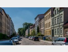 Eine gepflasterte Straßenansicht mit geparkten Autos auf beiden Seiten, flankiert von mehrstöckigen Wohnhäusern mit historischen Fassaden in verschiedenen Pastellfarben; im Hintergrund befinden sich Bäume unter einem klaren, blauen Himmel.