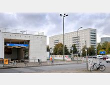 U-Bahn-Station Schillingstraße mit weiß gefliestem Eingang und blauer Beschilderung, im Hintergrund ein mehrstöckiges Wohngebäude und Bäume. Fahrräder stehen am Geländer, und Autos fahren auf der Straße daneben.