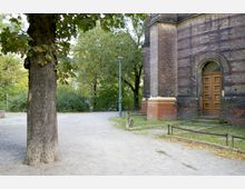 Ein sandiger Weg mit einem großen Baum im Vordergrund, umgeben von Grünflächen und Büschen. Rechts im Bild ist ein Teil eines Backsteingebäudes mit einer Holztür und einem runden Türbogen zu sehen, daneben eine grüne Laterne.