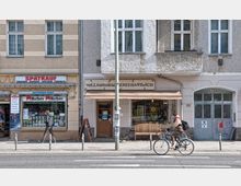 Straßenansicht mit einem Spätkaufgeschäft und einer Vollkornbäckerei in einem mehrstöckigen Wohngebäude. Vor den Geschäften sind Passanten unterwegs, ein Fahrradfahrer fährt auf der Straße, und ein hellbrauner Schirm sowie eine Holzbank befinden sich vor der Bäckerei.