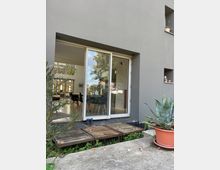 Moderne Hausfassade mit einer großen Glas-Schiebetür, die in einen Essbereich mit Tisch und Stühlen führt. Vor der Tür befindet sich eine kleine Holzplattform mit Pflanzen in Töpfen neben der Fassade.