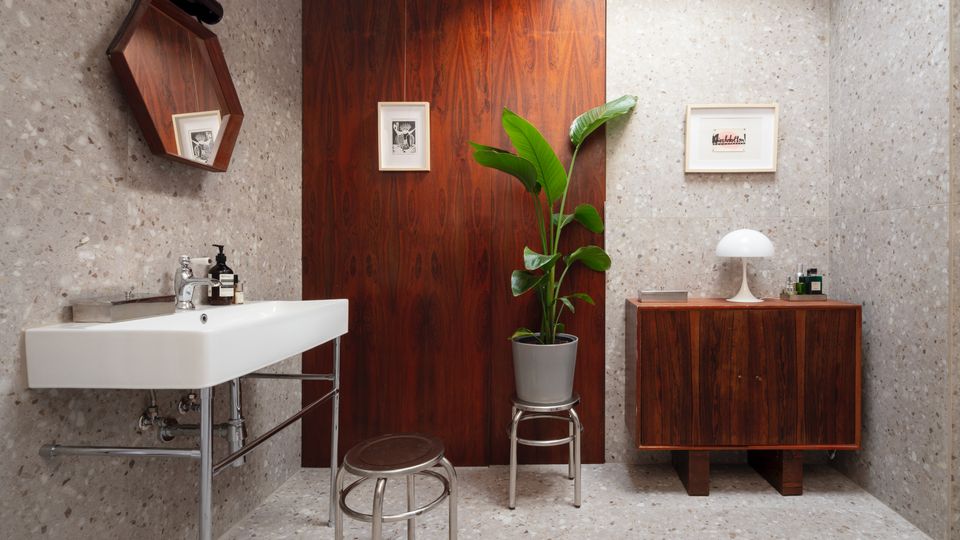 Modern bathroom with terrazzo walls and floor, a white wall-mounted sink, and a central wood-paneled partition. A skylight brightens the space, which includes a wooden cabinet with a table lamp, framed artwork, a round stool, and a potted plant.