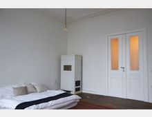 Ein minimalistisch eingerichtetes Zimmer mit weiß gestrichenen Wänden, einem niedrigen Bett mit weißer Bettwäsche und Kissen auf der linken Seite. Rechts steht ein weiß gekachelter Kaminofen vor einer Doppelflügeltür mit Milchglasscheiben, darüber hängt eine einzelne Glühbirne an einem Kabel.