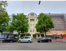 Wohnhaus in einer städtischen Straße mit heller Fassade und symmetrischen Fenstern, flankiert von zwei großen Bäumen. Links und rechts sind angrenzende Gebäude sichtbar, darunter ein Gerüst mit Bauarbeiten. Autos parken entlang der Straße.
