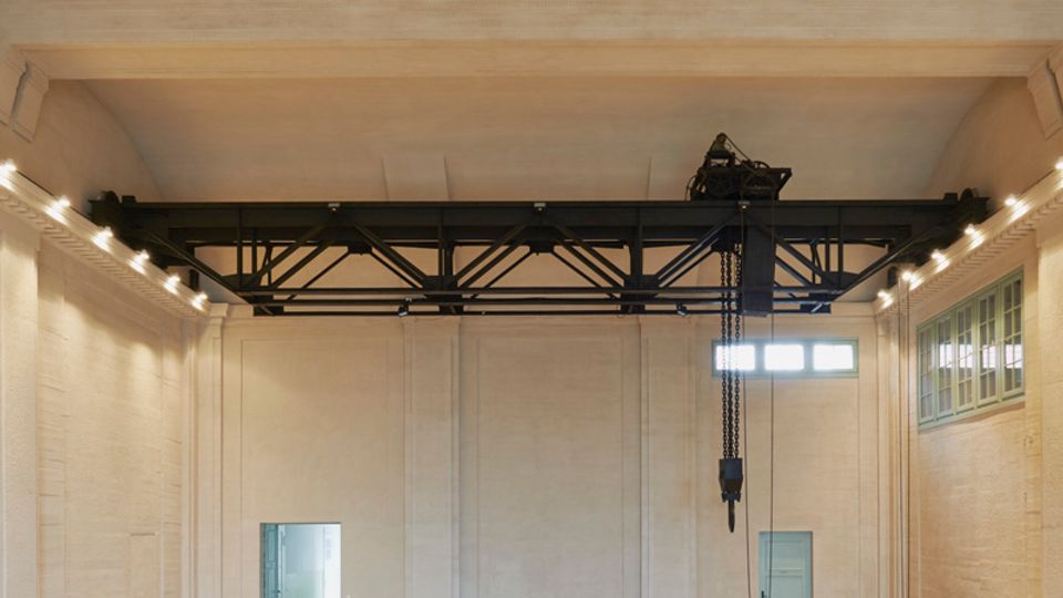 Großer, leerer Innenraum mit heller Kachelbodenfläche im Schachbrettmuster und einer hohen Kassettendecke mit dekorativen Öffnungen. An den Wänden befinden sich große Glasfenster sowie eine Metallkonstruktion mit Kette, die auf industrielle Nutzung hinweist.