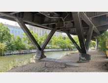 Blick unter eine Stahlbrücke mit dichtgefügten Trägern, die an einem sandigen Ufer eines Kanals steht. Im Hintergrund sind mit Graffiti verzierte Ufermauern, grüne Bäume und ein mehrstöckiges Gebäude mit blauer Fassade zu sehen.