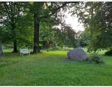 Ein Park mit grüner Wiese und großen Bäumen, darunter eine weiße Bank mit einem kleinen Tisch. Rechts im Vordergrund liegt ein großer Stein, und im Hintergrund steht eine rote Skulptur.