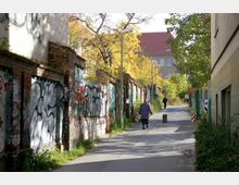 Schmaler Weg in einem urbanen Gebiet, gesäumt von alten Backsteinmauern mit Graffiti. Rechts stehen moderne Gebäude und Bäume mit Herbstlaub, während im Hintergrund ein größeres Gebäude mit rotem Dach sichtbar ist.