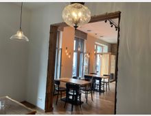 Ein gemütlicher Innenraum mit Holzböden, mehreren Holztischen und schwarzen Stühlen. Der Raum ist durch hängende Glühbirnen beleuchtet, hat große Fenster zur Straße hin und einen offenen Durchgang mit sichtbaren Holzrahmen.