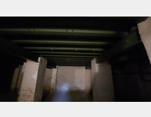 Dunkler Kellerraum mit weißen Backsteinwänden und einer Decke aus dunkelgrünen Holzbalken. Der Raum ist schlicht und enthält keine erkennbaren Einrichtungsgegenstände.