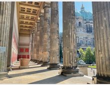 Blick durch die Säulengangarchitektur eines historischen Gebäudes mit dekorativen roten Wandfeldern und Kassettendecke, im Hintergrund der Berliner Dom mit grüner Kuppel und goldenen Details sowie Bäume und eine Statue davor.