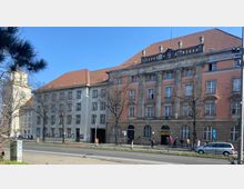 Historisches Gebäude mit pastellfarbenem Putz und roten Ziegeldächern, bestehend aus zwei Flügeln; der rechte Abschnitt ist mit Säulen und klassischen Ornamenten verziert. Im Vordergrund verlaufen eine Straße und ein Gehweg, Bäume ohne Laub stehen entlang der Fassade.