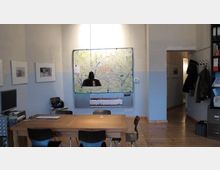 Ein schlicht eingerichteter Büroraum mit einem Holztisch und mehreren Stühlen. An der Wand hängt eine große Landkarte, darunter ein Regal mit Dokumenten, daneben befinden sich ein Computerarbeitsplatz und ein offener Zugang zu einem Flur.