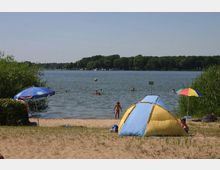 Ein Sandstrand mit Blick auf einen See, an dem Menschen schwimmen. Im Vordergrund stehen ein blau-gelbes Zelt und bunte Sonnenschirme, im Hintergrund Bäume am Ufer.