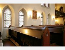 Innenraum einer Kirche mit hölzernen Sitzbänken, gotischen Spitzbogenfenstern aus buntem Glas und einem Liedanzeiger an der Wand. Die Wände sind hell gestrichen, und die Fenster lassen gefiltertes Tageslicht herein.