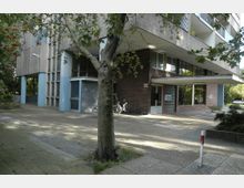 Mehrstöckiges Wohngebäude aus Beton mit braunen Klinkerwänden im Eingangsbereich, bodentiefen Fenstern und einem Vordach. Im Vordergrund steht ein Baum auf einer gepflasterten Fläche, umgeben von Grünflächen.