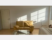 Wohnzimmer mit hellem Holzboden, einer gelben Ledercouch und einem Glastisch im Vordergrund. Eine weiße Tür, ein schlichter Stehlampe und eine minimalistische Wand mit Licht- und Schatteneffekten durch ein Fenster sind ebenfalls sichtbar.