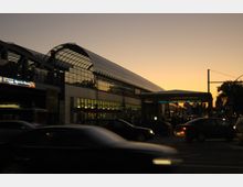 Ein moderner Bahnhof mit großflächigen Glasdächern und einer beleuchteten Fassade bei Sonnenuntergang, im Vordergrund fahren Autos auf einer belebten Straße. Links ist ein Eingang mit einem Schild und weiter rechts erkennbar ein U-Bahn-Zugang.