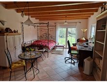 Ein gemütlich eingerichtetes Zimmer mit einem Metallbett, einem kleinen runden Esstisch mit Stühlen, einem Schreibtisch mit Bürostuhl und Regalen. Der Raum hat helle Wände, Holzbalkendecken und große Fenster mit Blick auf einen grünen Garten.