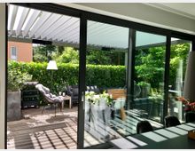 Moderne Terrasse mit Holzboden, sichtbarem Glas-Schiebetürzugang und einer Überdachung aus Lamellen. Die Terrasse ist mit Sitzmöbeln, Pflanzen, einem Beistelltisch und einer Hecke als Sichtschutz gestaltet, im Hintergrund ist dichtes Grün zu erkennen.