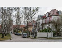 Wohnviertel mit einer gepflasterten Straße, gesäumt von weißen Birken und geparkten Autos. Rechts befinden sich Häuser mit roten Ziegeldächern und großen Fenstern, im Hintergrund ein Gebäude mit grünlicher Kuppel.