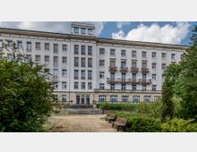 Fassade eines mehrstöckigen, historischen Wohngebäudes mit symmetrisch angeordneten Fenstern und kleinen Balkonen. Im Vordergrund sind ein kleiner Weg, Büsche, Bäume und zwei Holzbänke zu sehen.