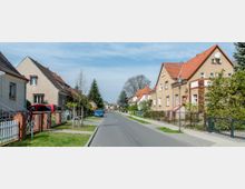 Straßenszene in einem Wohngebiet mit Reihen von Einfamilienhäusern mit roten Ziegeldächern und beige-braunen Fassaden. Die Straße ist von Gehwegen, Vorgärten und einigen parkenden Autos gesäumt, umgeben von Bäumen und Hecken.