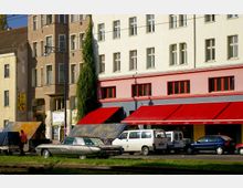 Straßenszene mit mehrstöckigen Wohn- und Geschäftshäusern, einem älteren Gebäude mit brauner Fassade und einem renovierten Gebäude mit heller Fassade und roten Markisen im Erdgeschoss. Im Vordergrund geparkte Autos, darunter ein silberner Oldtimer, sowie ein Gehweg mit einer grünen Rasenfläche.