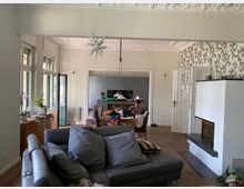 Wohnzimmer mit moderner Einrichtung, darunter ein graues Ledersofa, eine offene Küche im Hintergrund und dekorative Deckenstuckaturen. Große Fenster lassen Tageslicht herein, und ein Kamin sowie ein Esstisch mit Stühlen prägen den Raum.