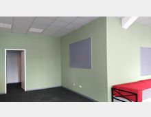 Innenraum eines einfachen Büros mit blassgrünen Wänden. Zu sehen sind eine offene Tür, eine Wand mit einer grauen Pinnwand sowie eine rote Couch im rechten Bildbereich.