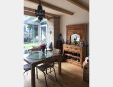 Helles Wohnzimmer mit großen gläsernen Terrassentüren, einem Holztisch mit Glasplatte und Stühlen mit Rattanoptik. Rechts steht eine geschnitzte Holzkommode mit dekorativen Elementen und Weinflaschenfächern, daneben einzelne Musikinstrumente und ein Buddha-Statue-Dekor. Im Hintergrund ist ein Garten sichtbar.