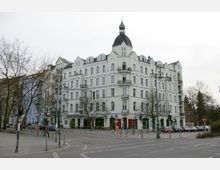 Eckgebäude in heller Fassade mit historischer Architektur und einem Turm mit Kuppel, umgeben von Bäumen und Straßenlaternen. Im Erdgeschoss befinden sich Geschäfte, während auf der Straße geparkte Autos und eine Kreuzung sichtbar sind.