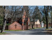 Straßenszene mit einem breiten Gehweg und Bäumen, links eine rote Backsteinmauer mit einem Kreuz und dahinter eine Backsteinkirche mit Satteldach. Rechts im Bild befinden sich helle Gebäude mit Ziegelmauern und leicht abgerundeten Fenstern, umgeben von Bäumen.