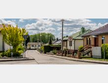 Wohngebiet mit Einfamilienhäusern, von denen einige grüne oder rote Dächer haben, an einer asphaltierten Straße mit Tempo-30-Zone. Links und rechts der Straße sind Gärten mit Bäumen und Sträuchern, im Hintergrund ein Wald unter einem leicht bewölkten Himmel.