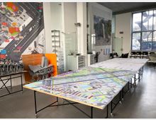 Ein moderner Arbeitsraum mit großen Tischen, auf denen Stadtpläne und Modelle liegen. Die Wände sind mit großformatigen Karten und Illustrationen dekoriert, und im Hintergrund sind Regale, Stühle und große Fenster zu sehen.