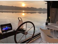 Innenraum eines modernen Hausbootes mit Blick durch ein großes Fenster auf einen ruhigen See bei Sonnenuntergang. Im Vordergrund ein Steuerrad, ein kleiner Tisch mit weißen Stühlen und eine minimalistische Dekoration.