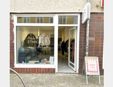 Schaufenster und Eingang eines kleinen Bekleidungsgeschäfts mit der Aufschrift „ALL FAB ETHICAL“, dahinter Inneneinrichtung mit Kleidung auf Kleiderständern und Sitzmöbeln. Daneben steht ein Aufsteller mit Werbung für einen „Sample Sale“.